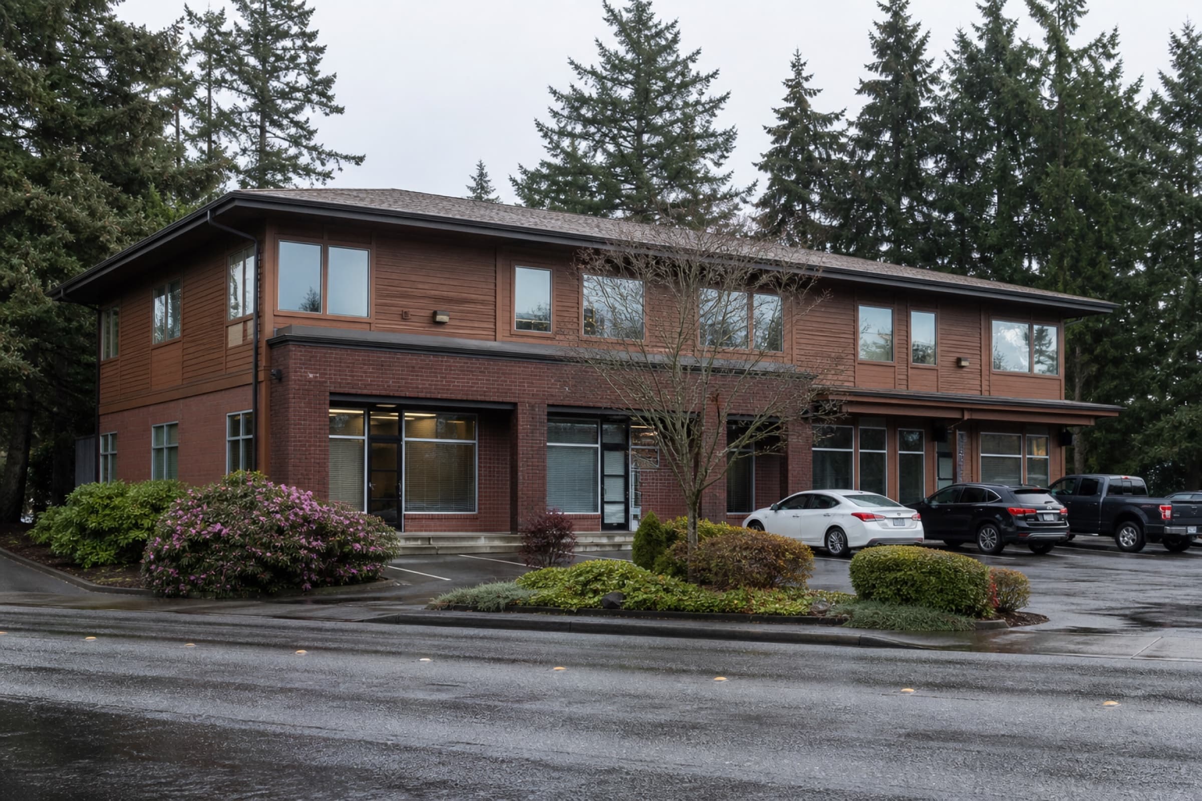 A two-story brick and cedar commercial building with ground-floor shops and evergreens behind it